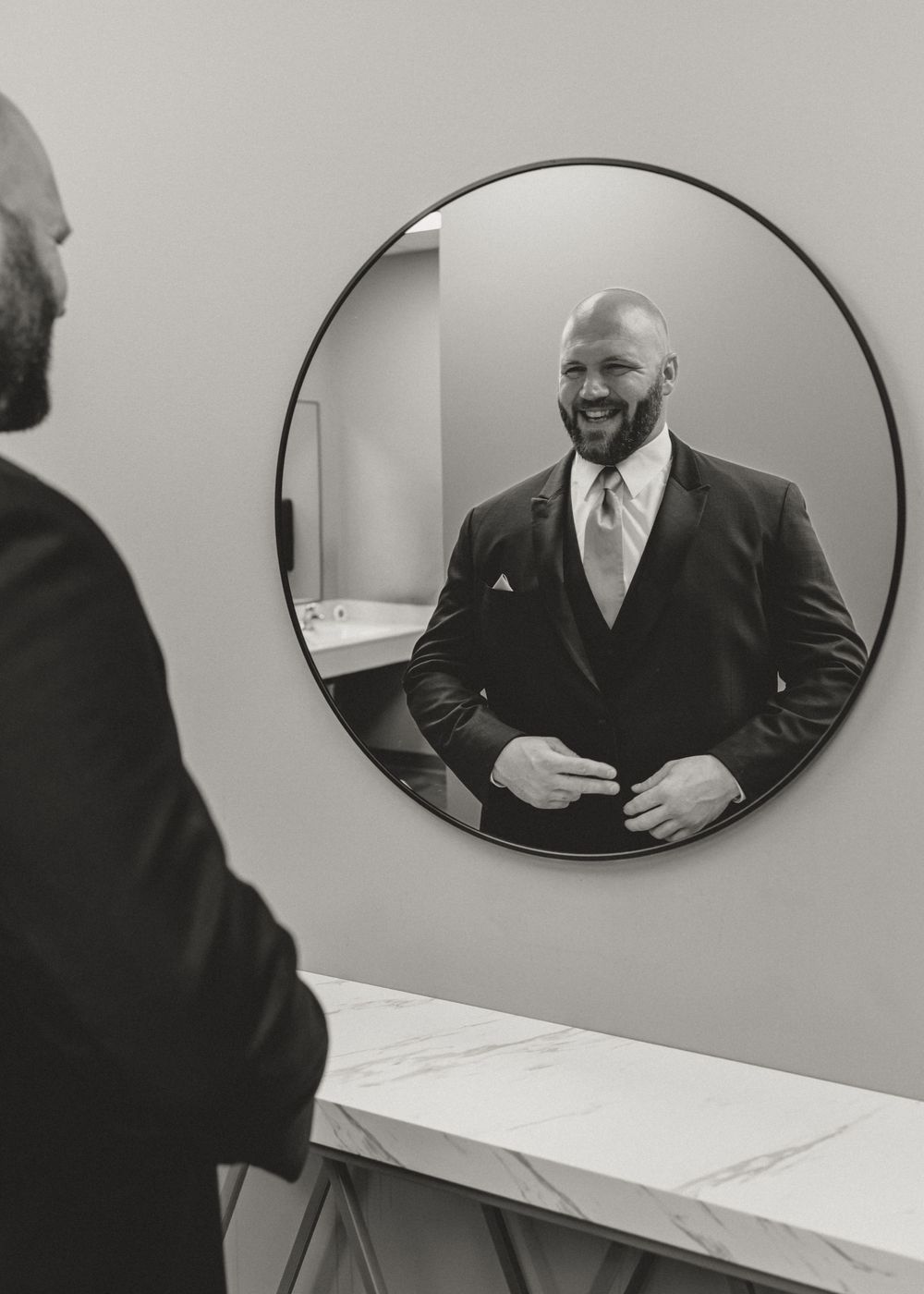 A smiling man in a dark suit adjusts his jacket while looking at his reflection in a round mirror.