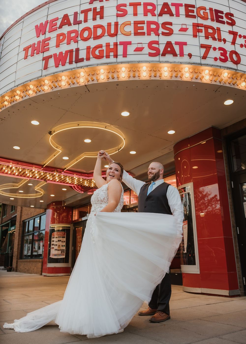 Bride and groom dancing joyfully outside a vintage theater marquee in wedding attire.