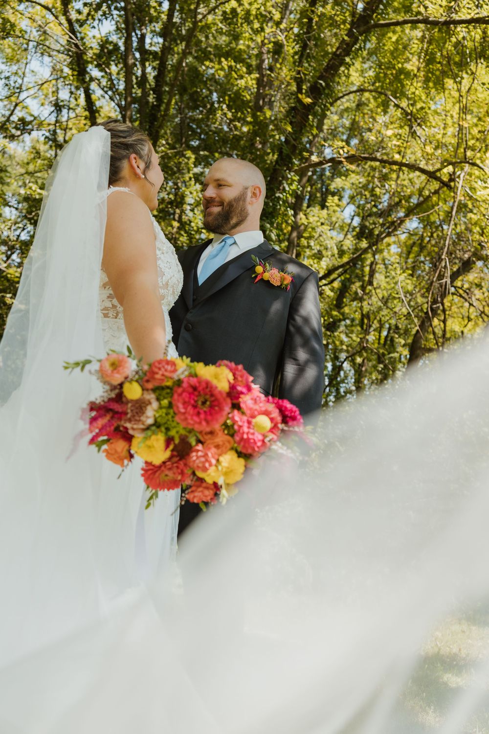 Bride and groom share a romantic moment outdoors, with a flowing veil and vibrant pink and yellow floral bouquet.