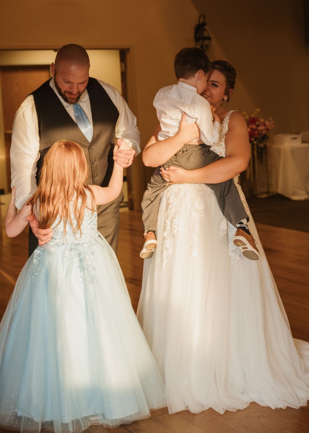 Bride and groom dancing with children at wedding reception, flower girl in blue dress, warm indoor venue.