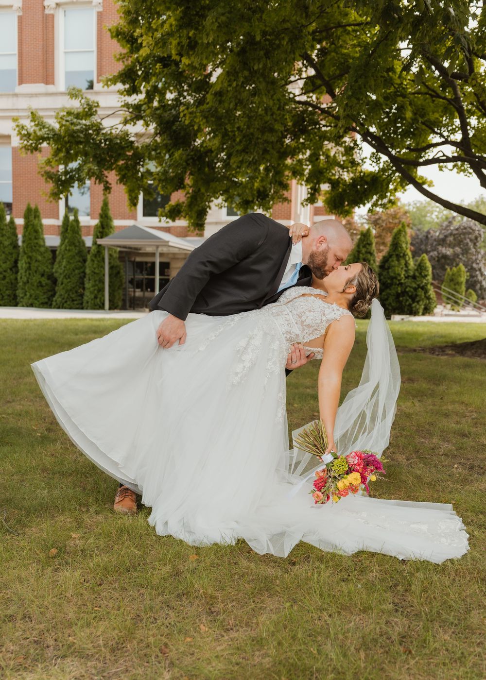 Groom dips bride for a romantic kiss outdoors, her white lace gown and veil flowing, holding a colorful bouquet near a brick building.