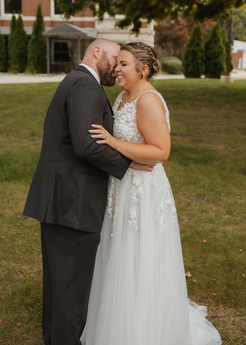 Bride in floral lace gown and groom in gray suit share a romantic moment outdoors on their wedding day.