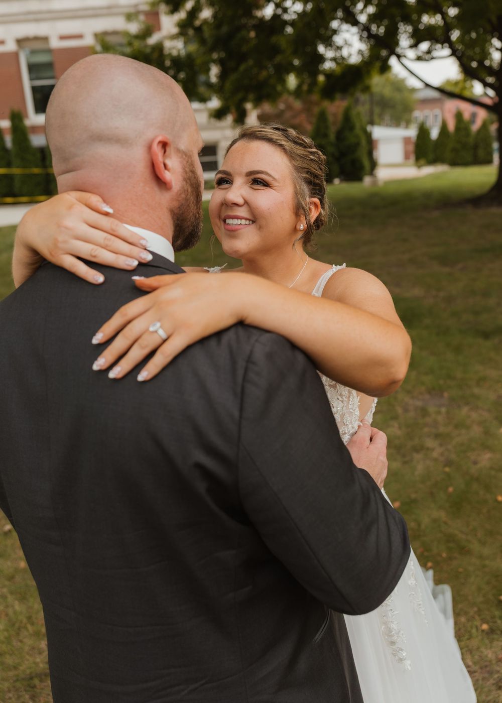 Bride in white gown embraces groom in dark suit outdoors, smiling joyfully, showcasing her diamond engagement ring.