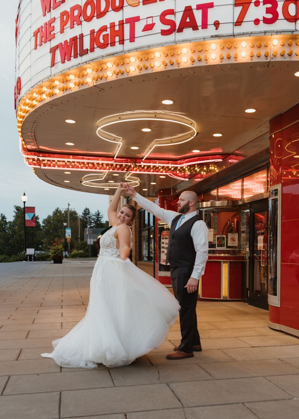 Bride and groom dancing in front of a lit theater marquee at twilight, bride in white gown, groom in gray vest.