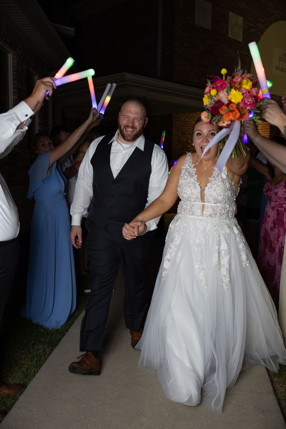 Bride and groom exit wedding reception through glowing foam stick tunnel held by guests at night.
