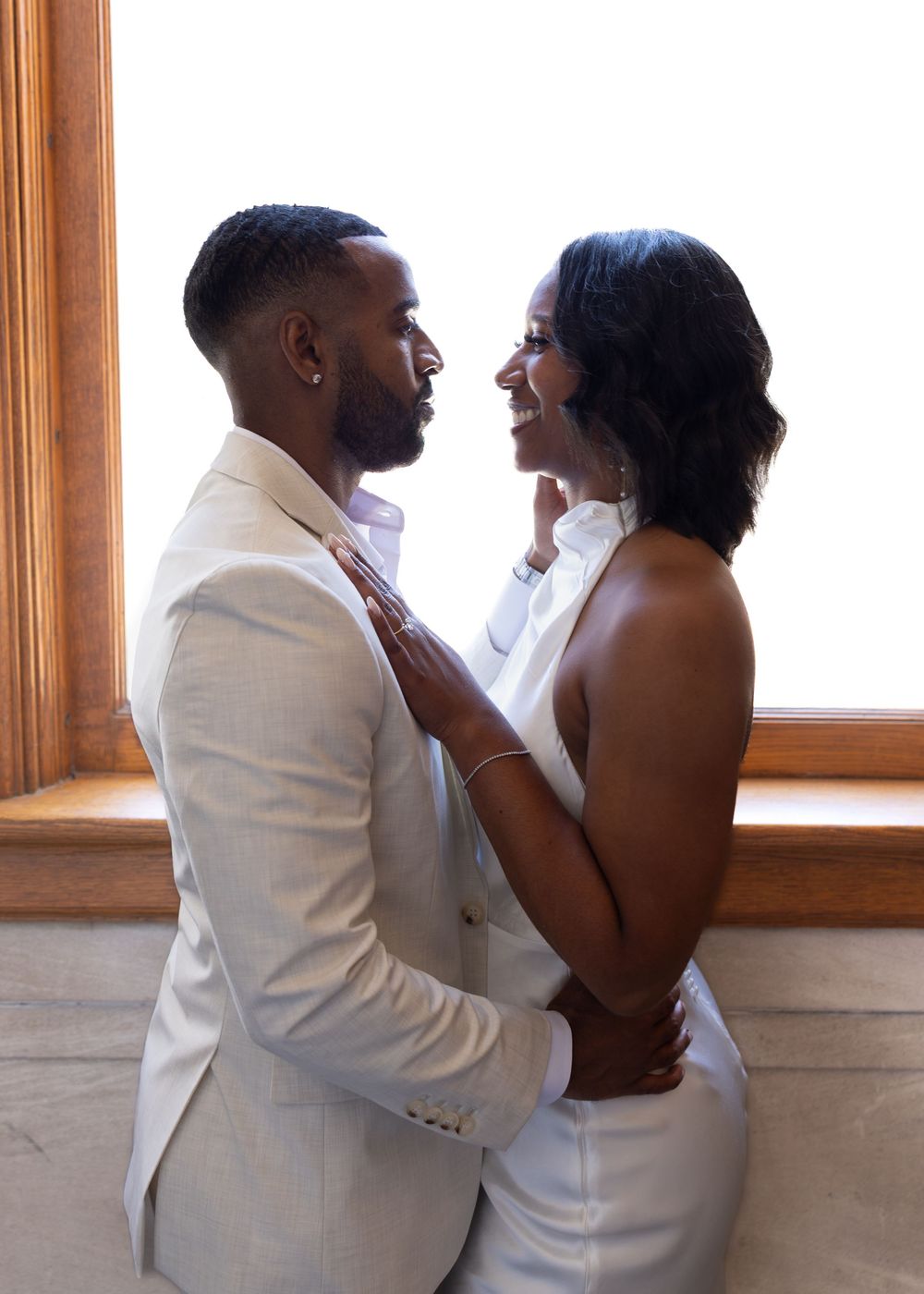 Elegant couple in white attire sharing a romantic moment by a bright window.