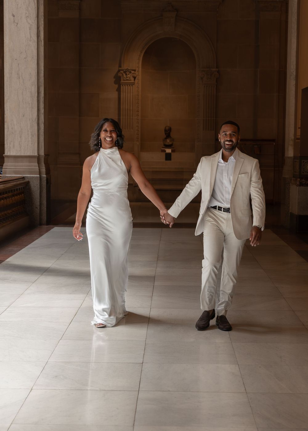 Joyful newlywed couple in white wedding attire walking hand in hand through grand marble hall.