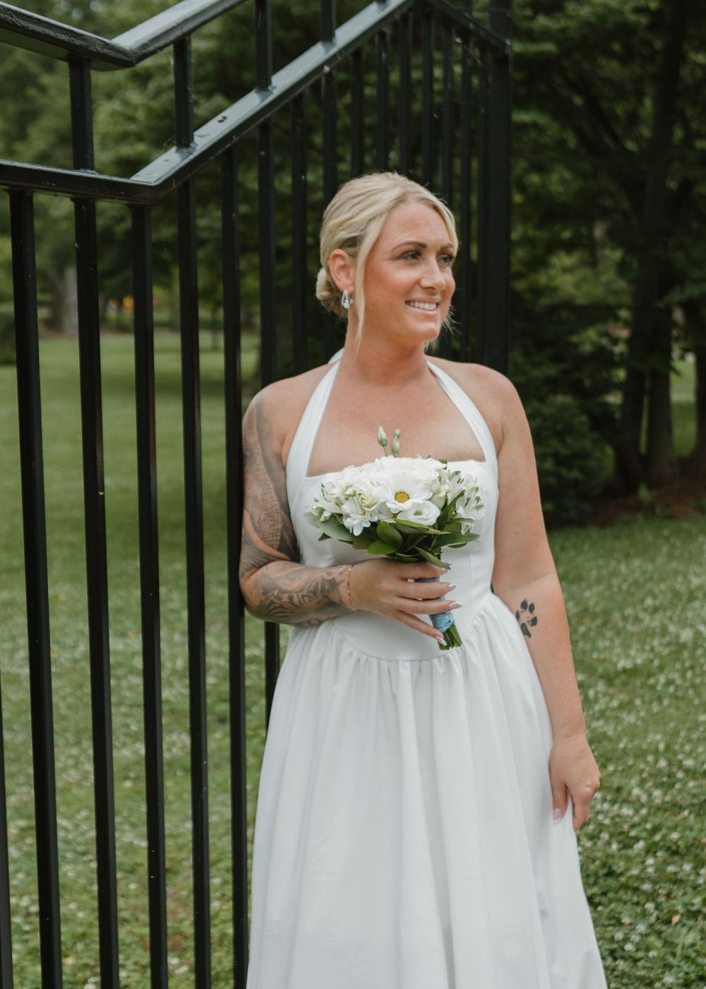 Tattooed bride in white halter dress holding daisy bouquet near black iron gate outdoors.