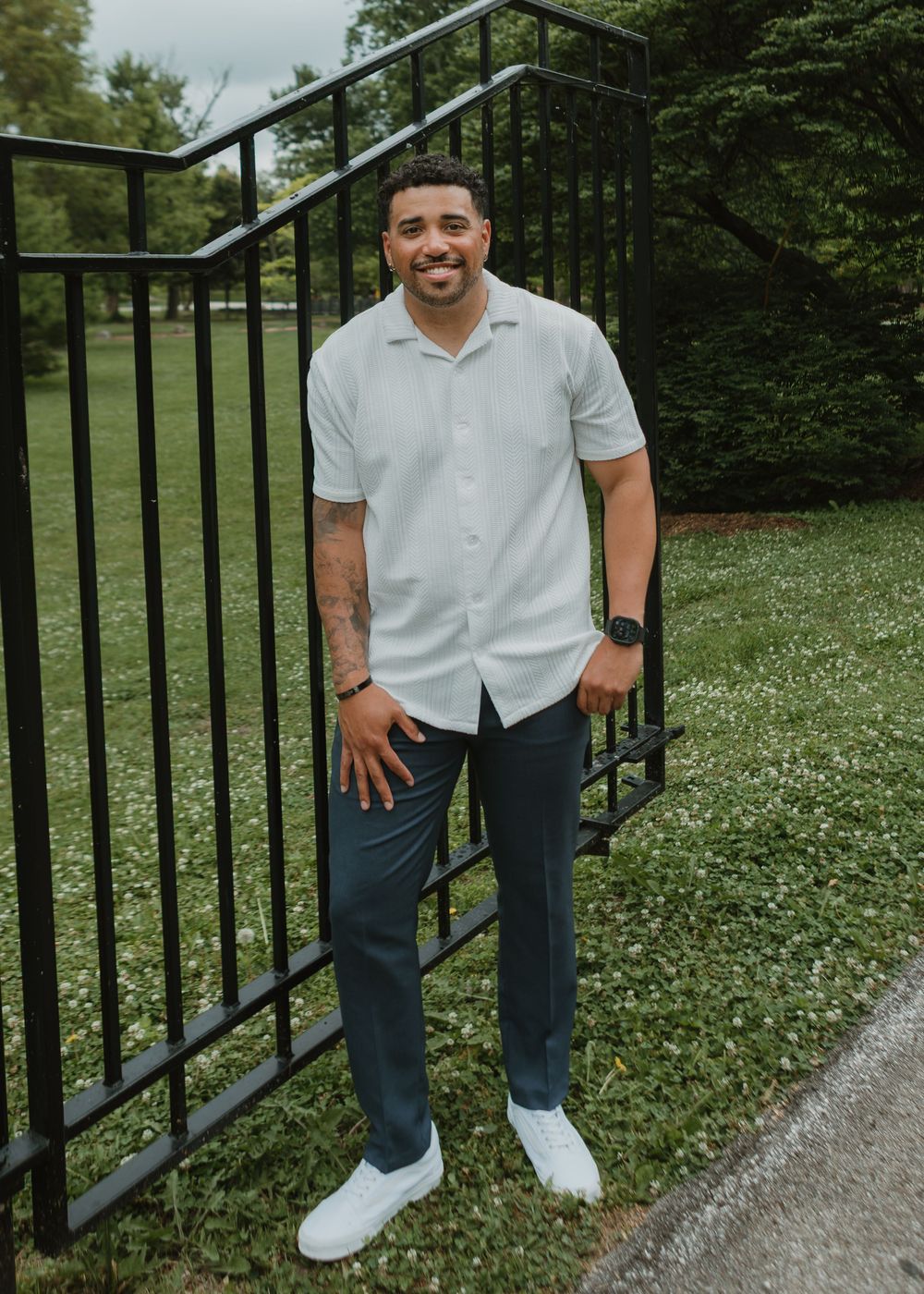 Smiling groom in light gray shirt and dark pants posing by black iron gate in park.