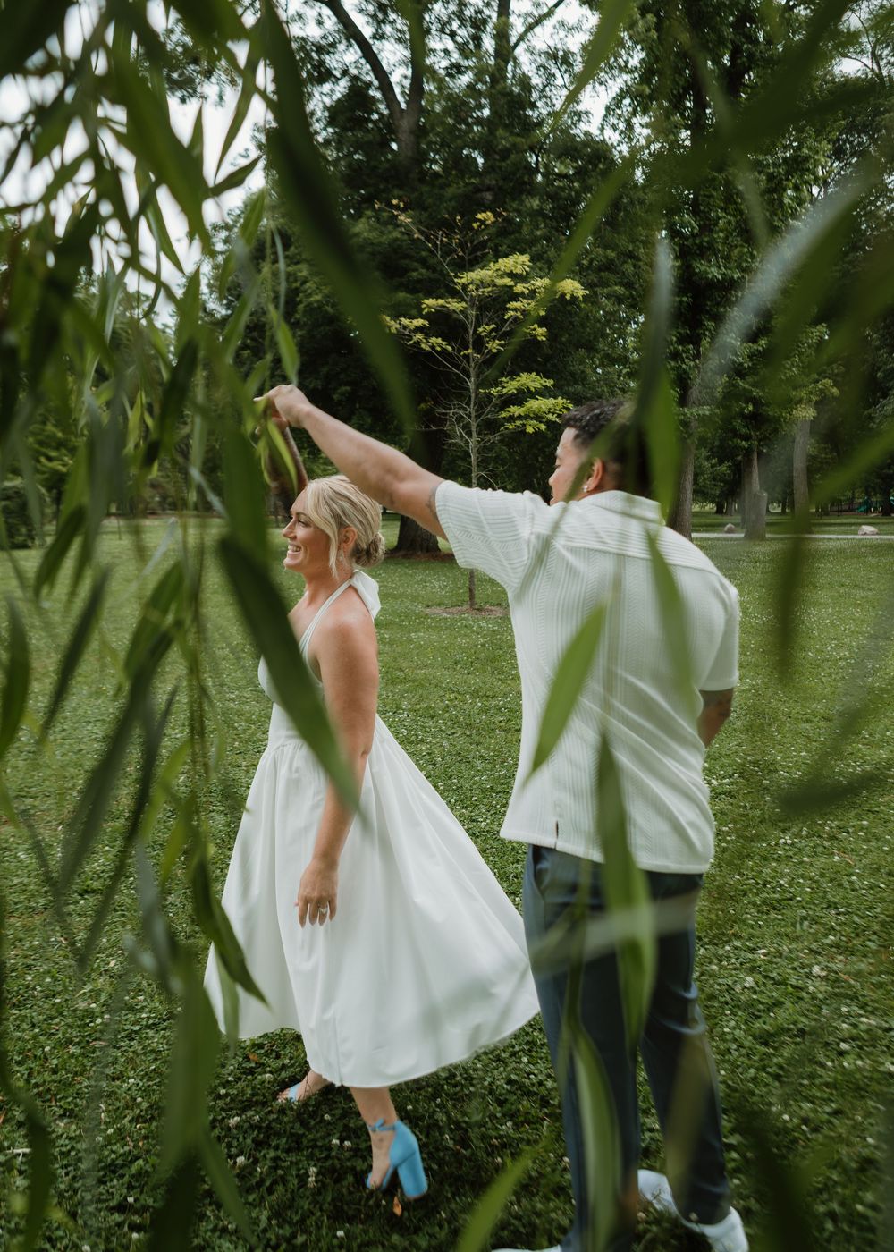 Couple dancing outdoors in a lush green park, man spinning woman in white dress and blue heels.