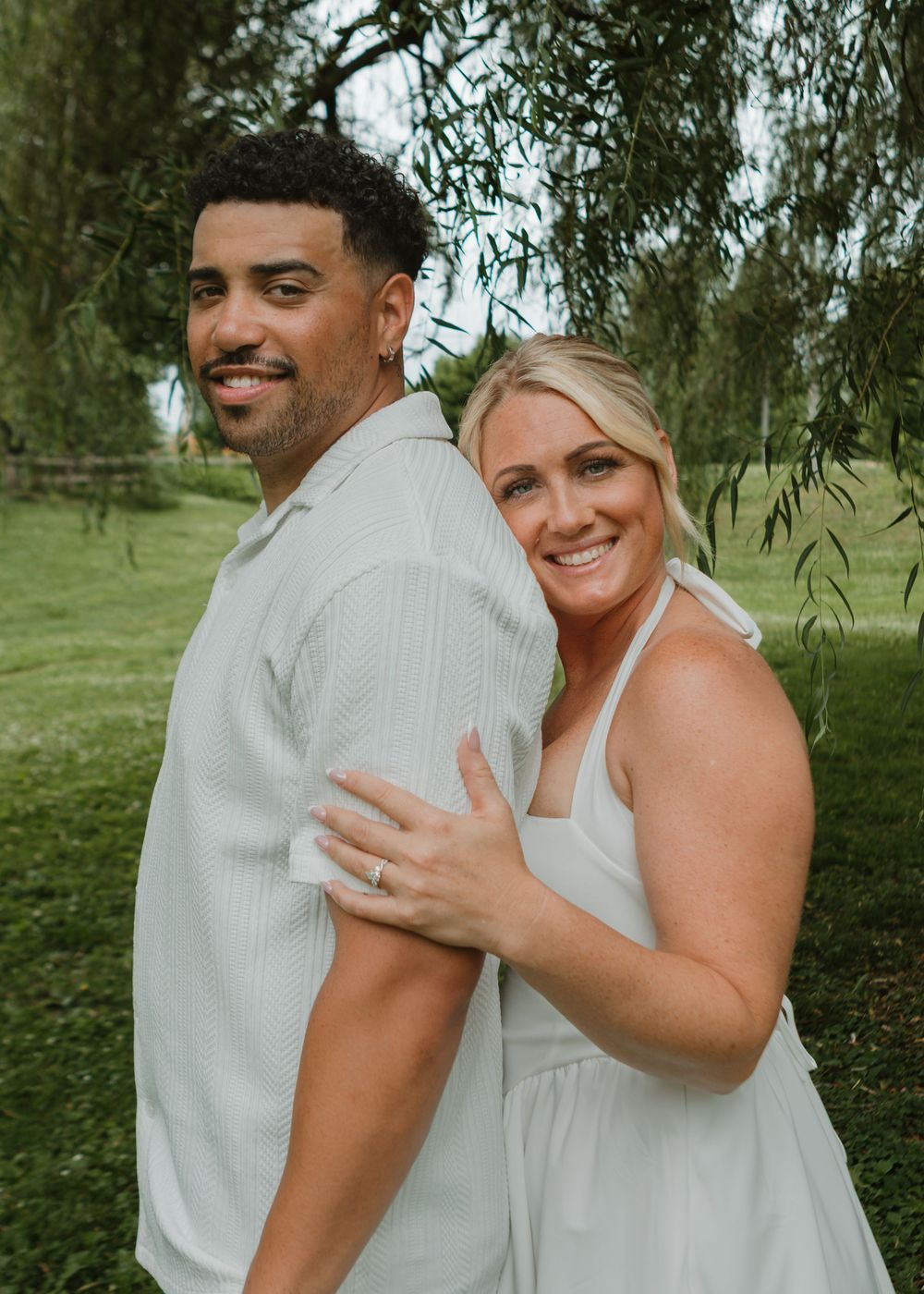 Happy interracial couple embracing outdoors under willow tree in matching white outfits.