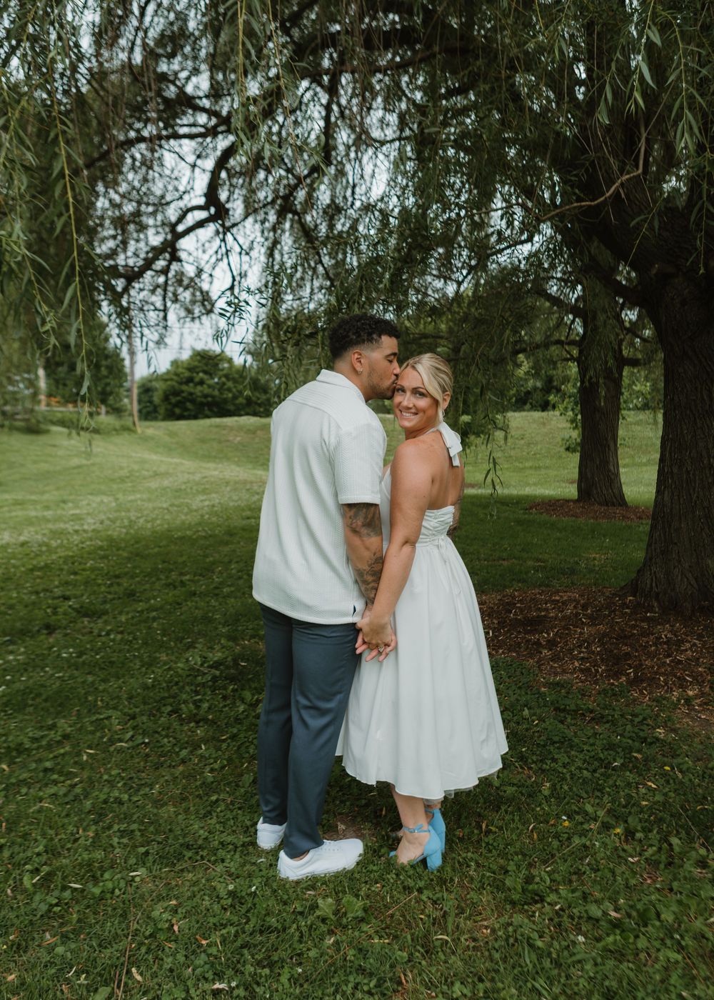 Romantic couple embracing under a weeping willow tree, woman in white midi dress and blue heels.