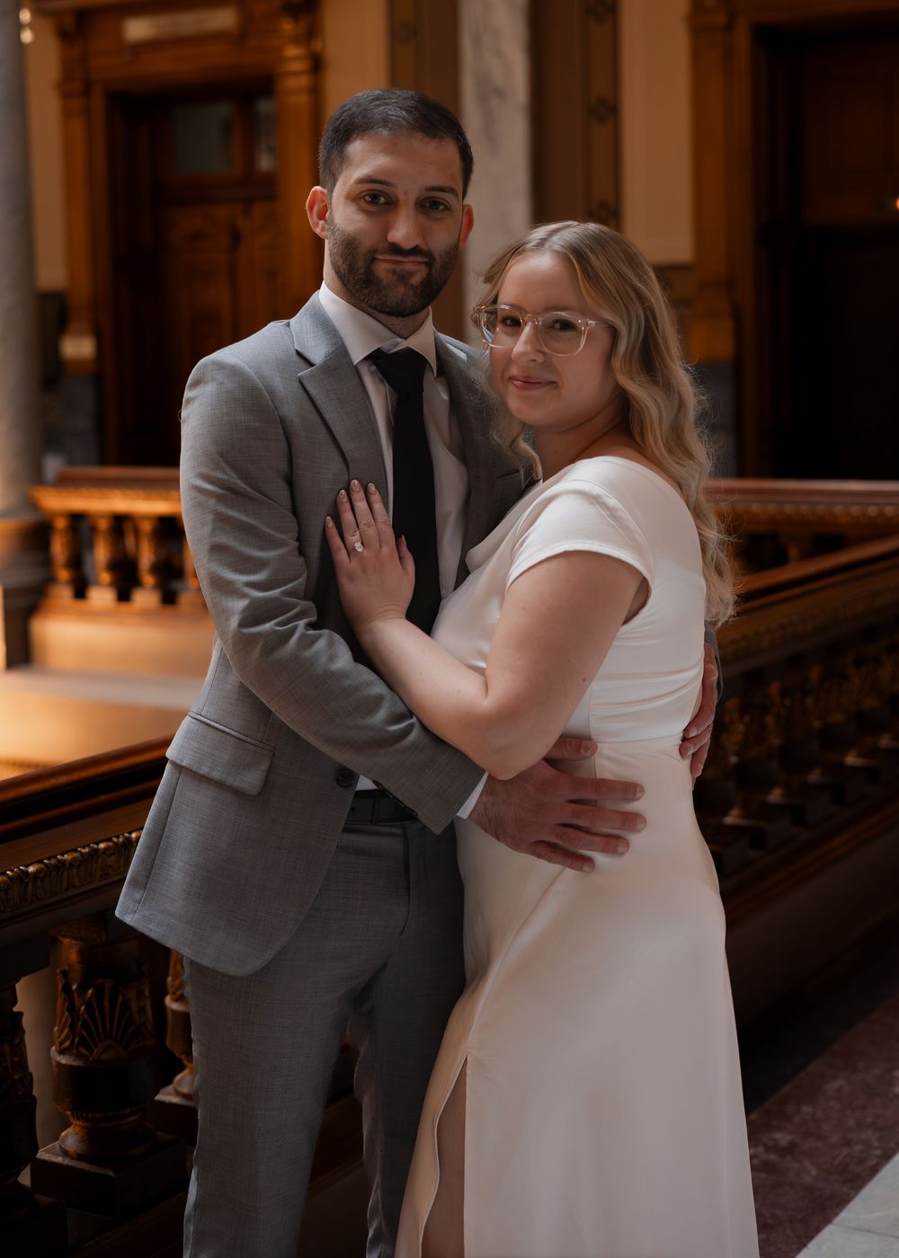 Elegant couple posing in a grand hall, man in gray suit and woman in white wedding dress with glasses.