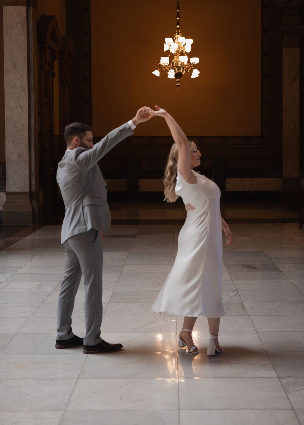 Couple sharing a romantic dance in an ornate marble hall beneath a glowing chandelier.