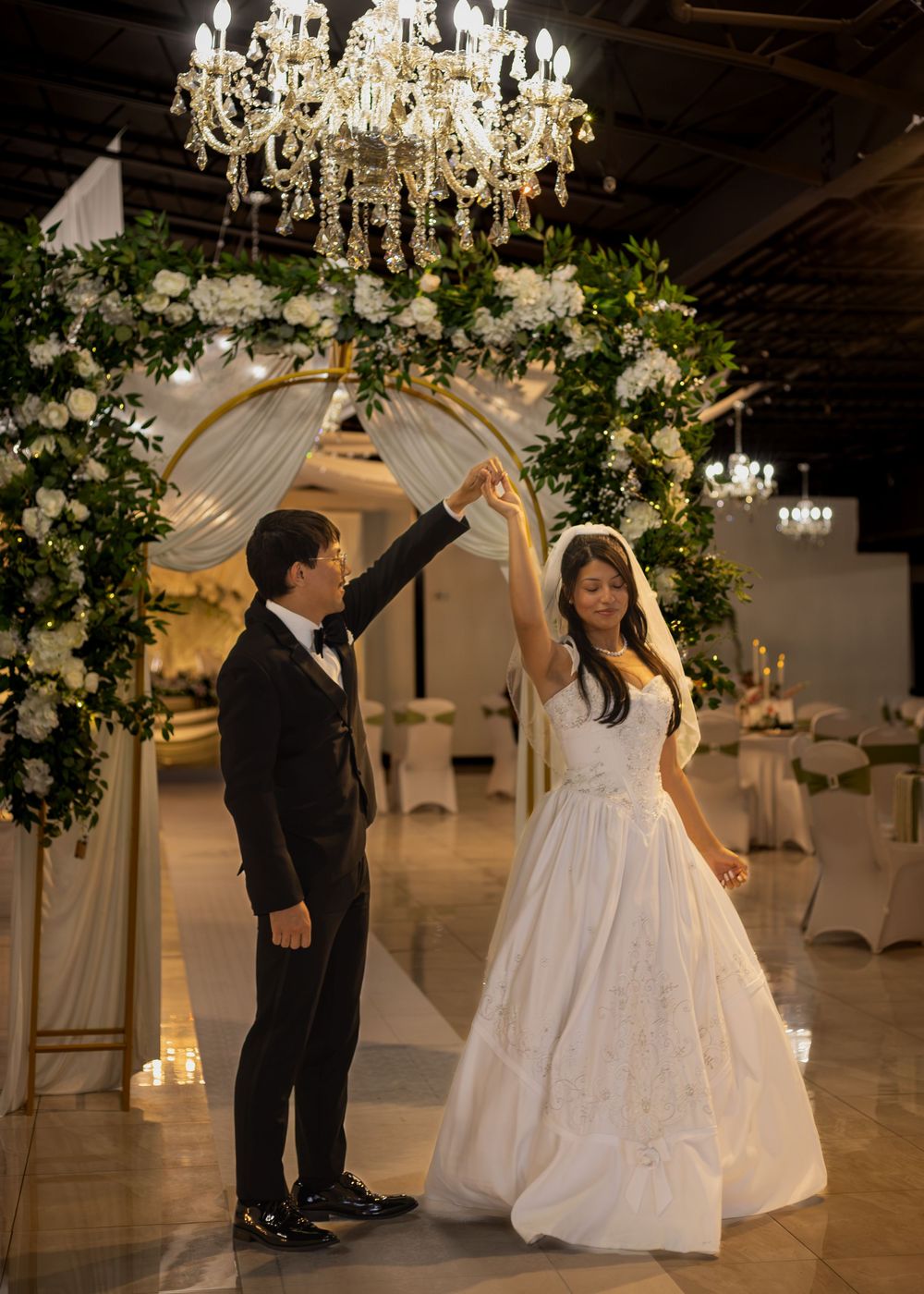 Bride and groom sharing first dance under chandelier and floral arch at wedding reception.