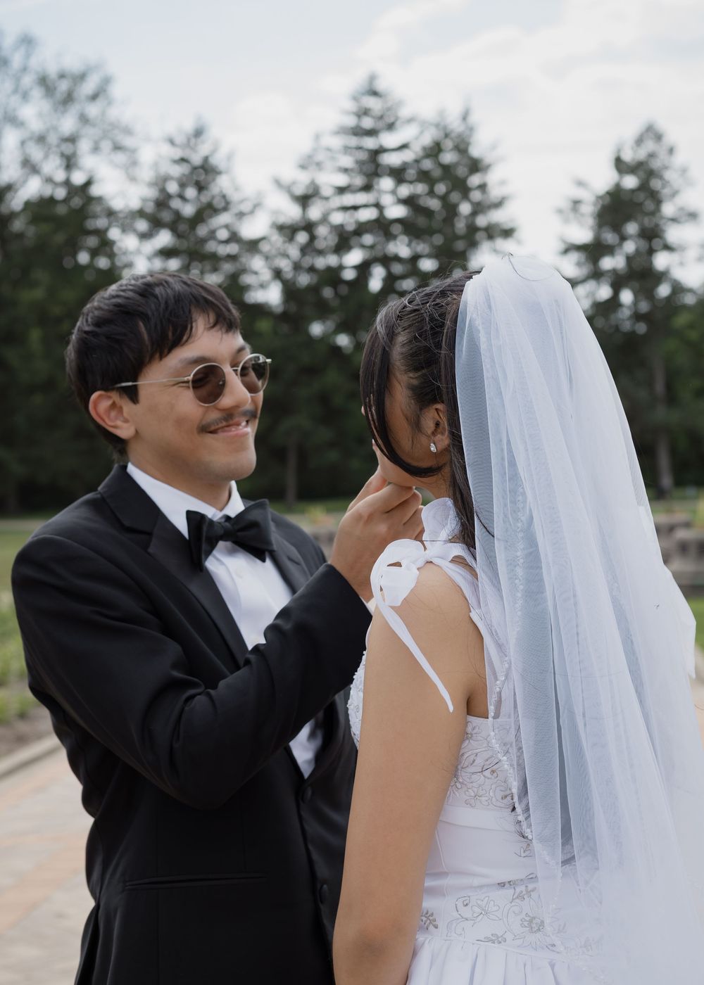 Smiling groom in black tuxedo adjusting bride's veil outdoors with trees in background.