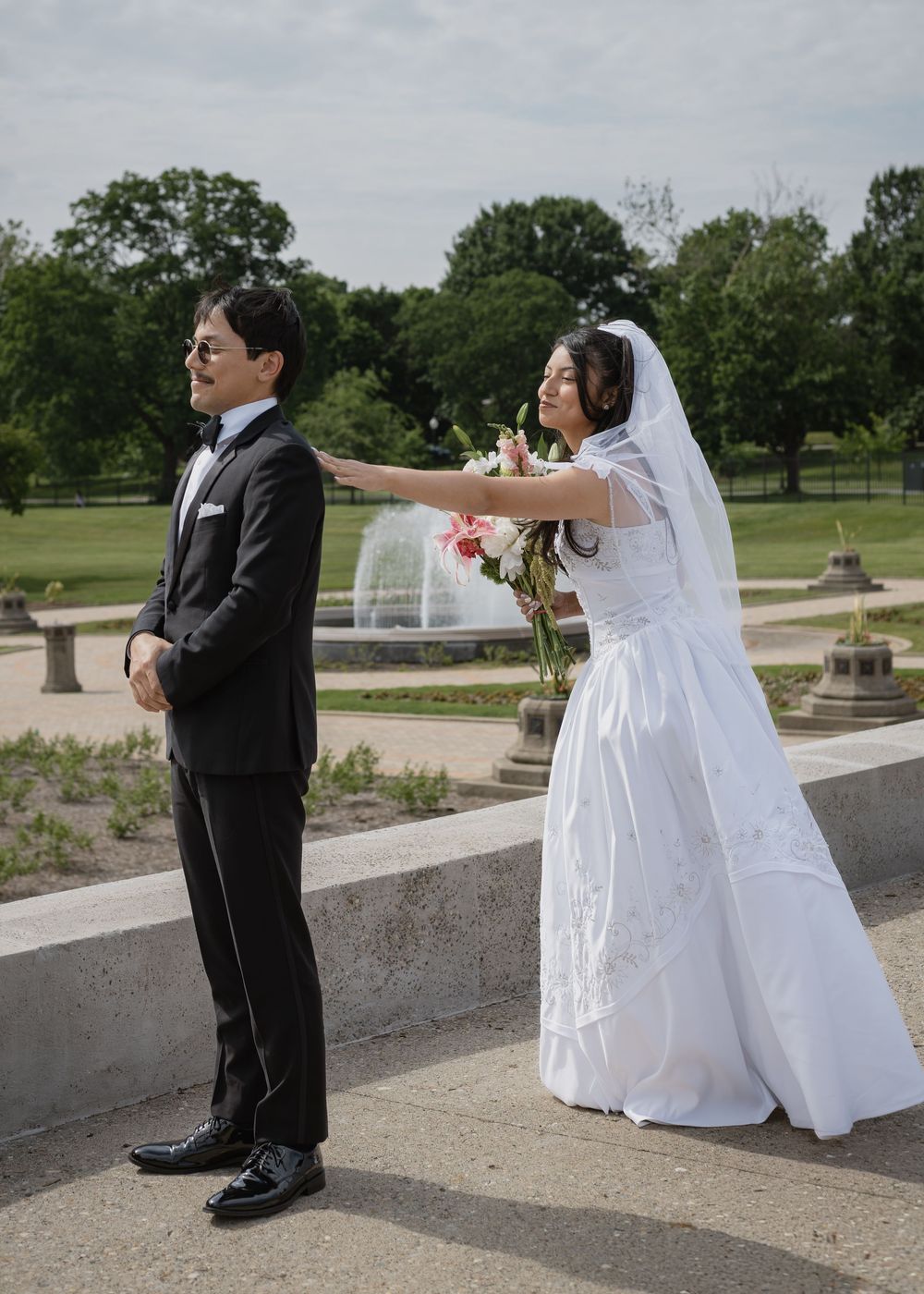 Bride in white gown tapping groom's shoulder during outdoor first look near fountain.