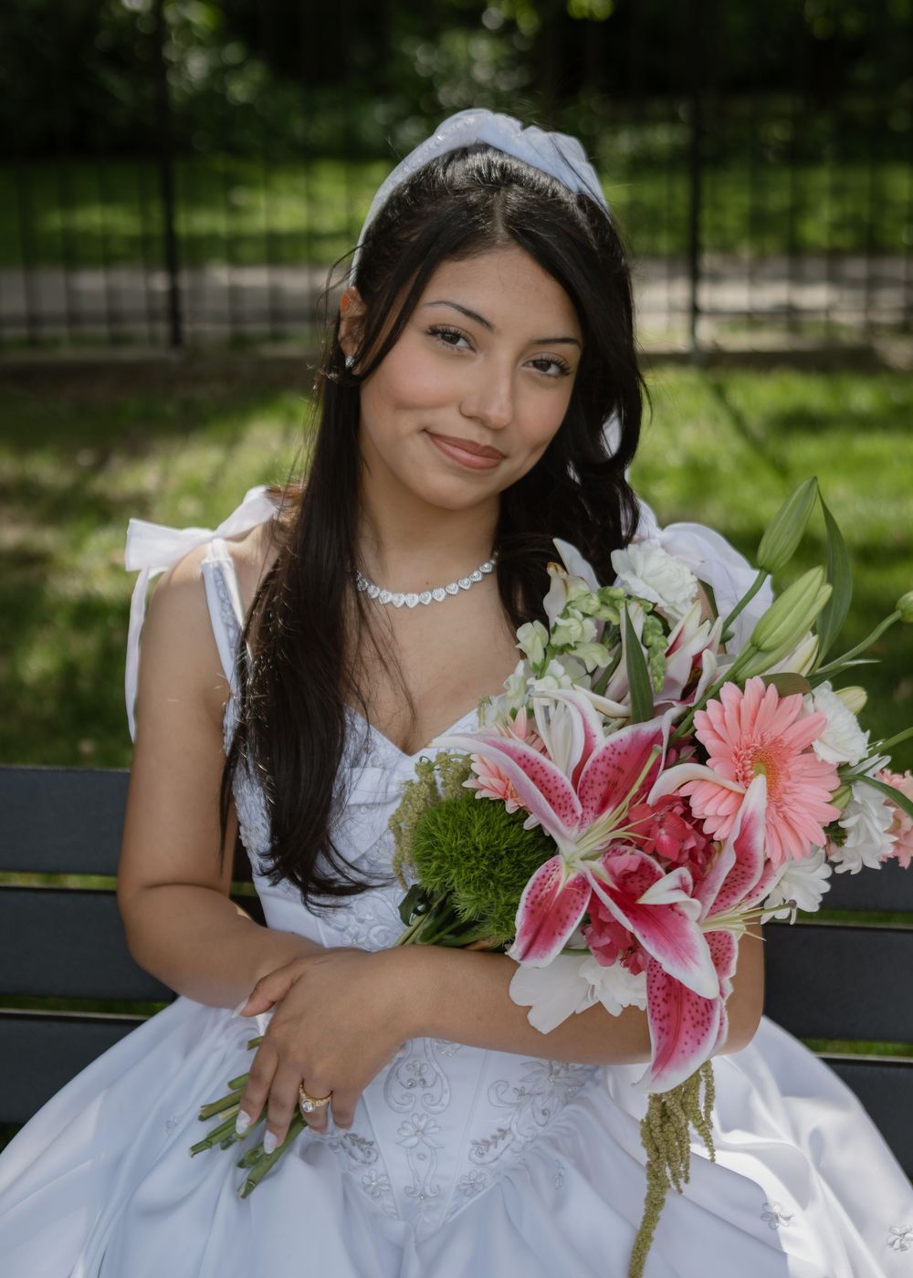 Beautiful bride in white gown holding pink and white floral bouquet seated in a park.