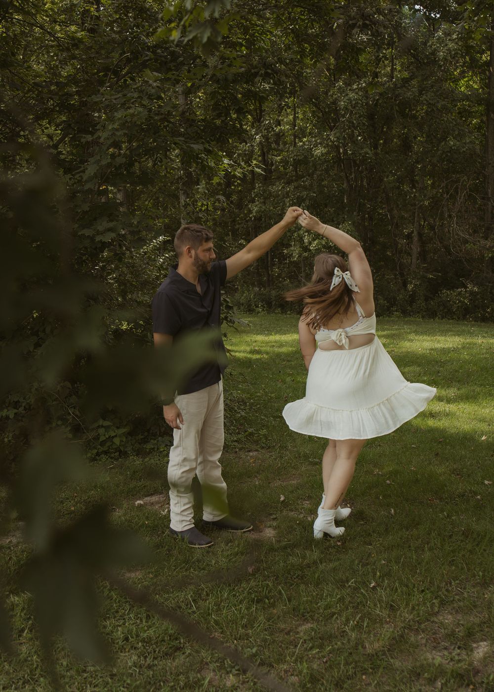 Couple dancing outdoors in a lush green forest, woman in white dress being twirled by man in dark shirt.