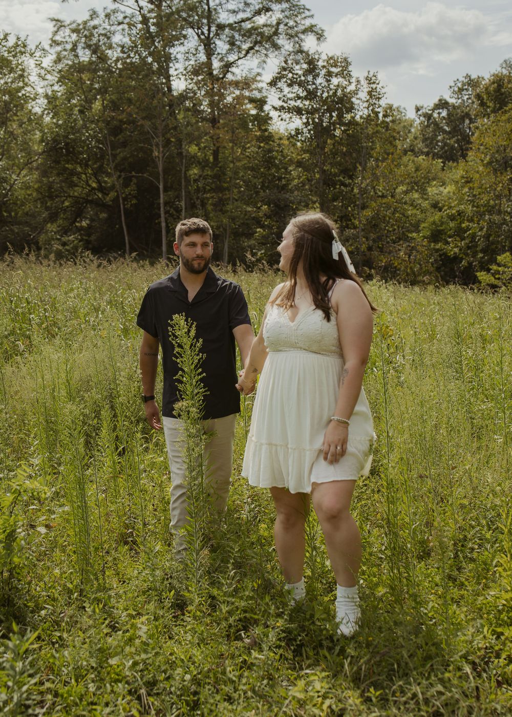 Couple holding hands in a lush green meadow, woman in white dress, man in black shirt, trees in background.
