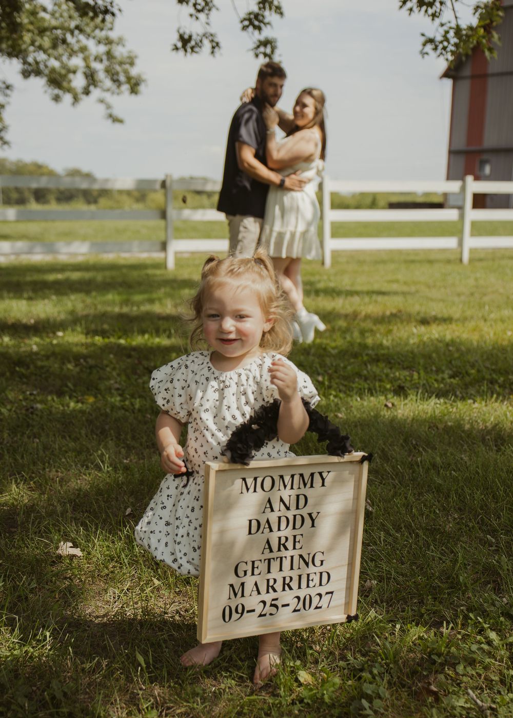 Toddler girl holds a wooden sign announcing parents wedding date 09-25-2027 while couple embraces on a farm.