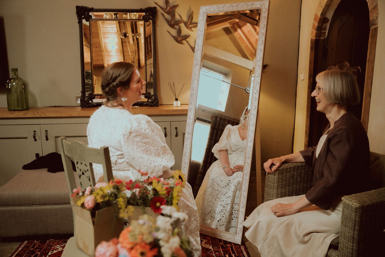 Bride in white lace gown views herself in tall mirror while a seated woman watches in a rustic barn setting.