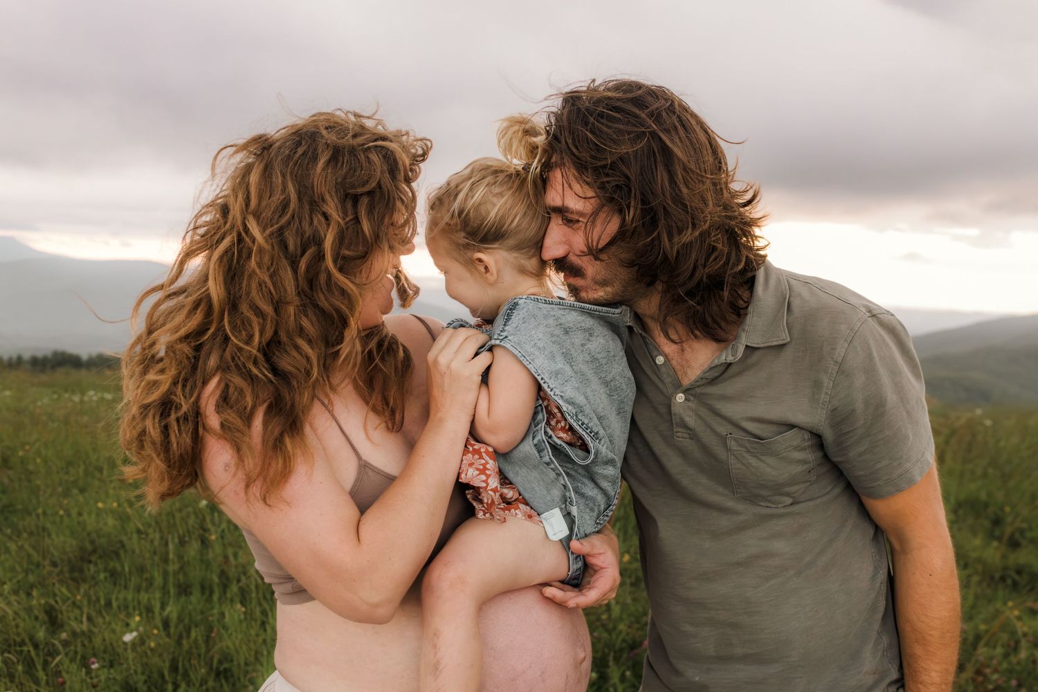 Family of three sharing a warm embrace under a cloudy sky, surrounded by lush greenery.