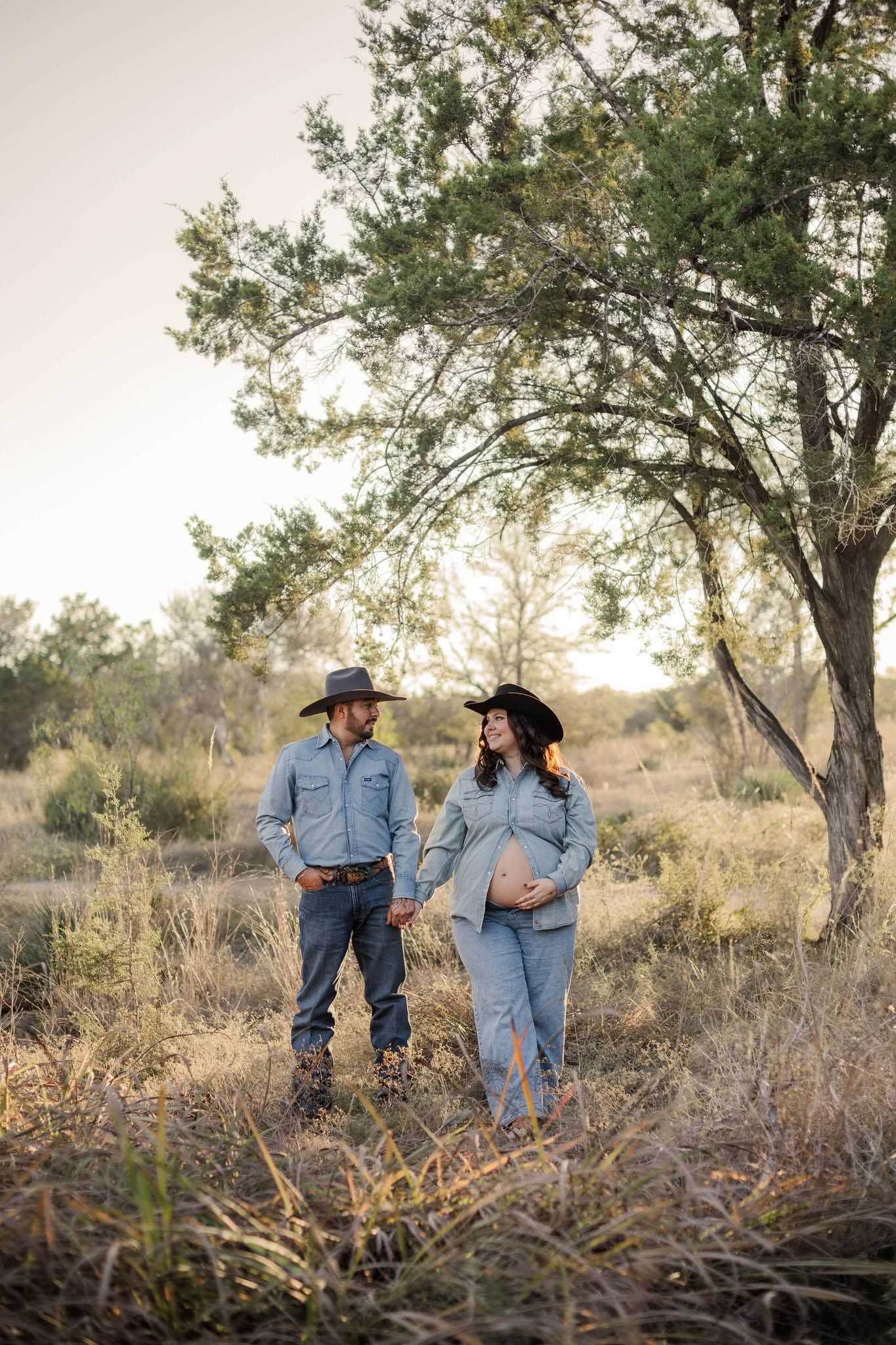 Expecting couple in matching denim and cowboy hats pose outdoors in a rustic field during a golden hour maternity photo session.