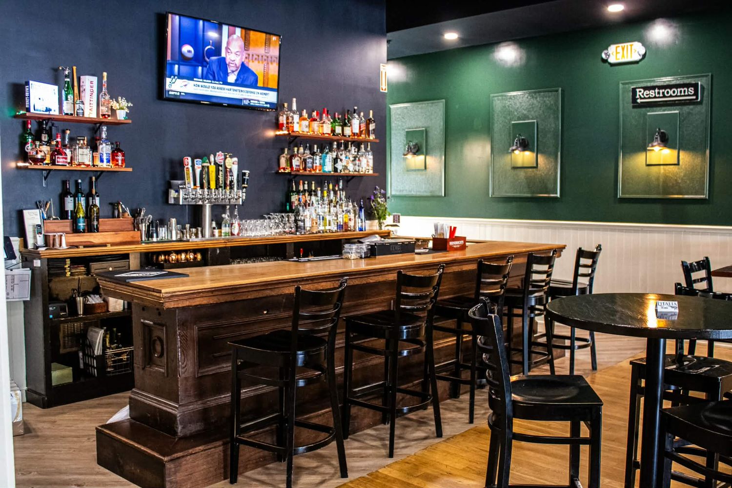 Cozy bar interior with wooden counter, black stools, wall-mounted TV, and shelves of liquor bottles in a green-accented space.