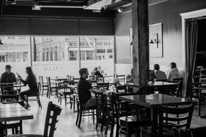 Black and white photo of a casual restaurant interior with patrons dining near large windows.