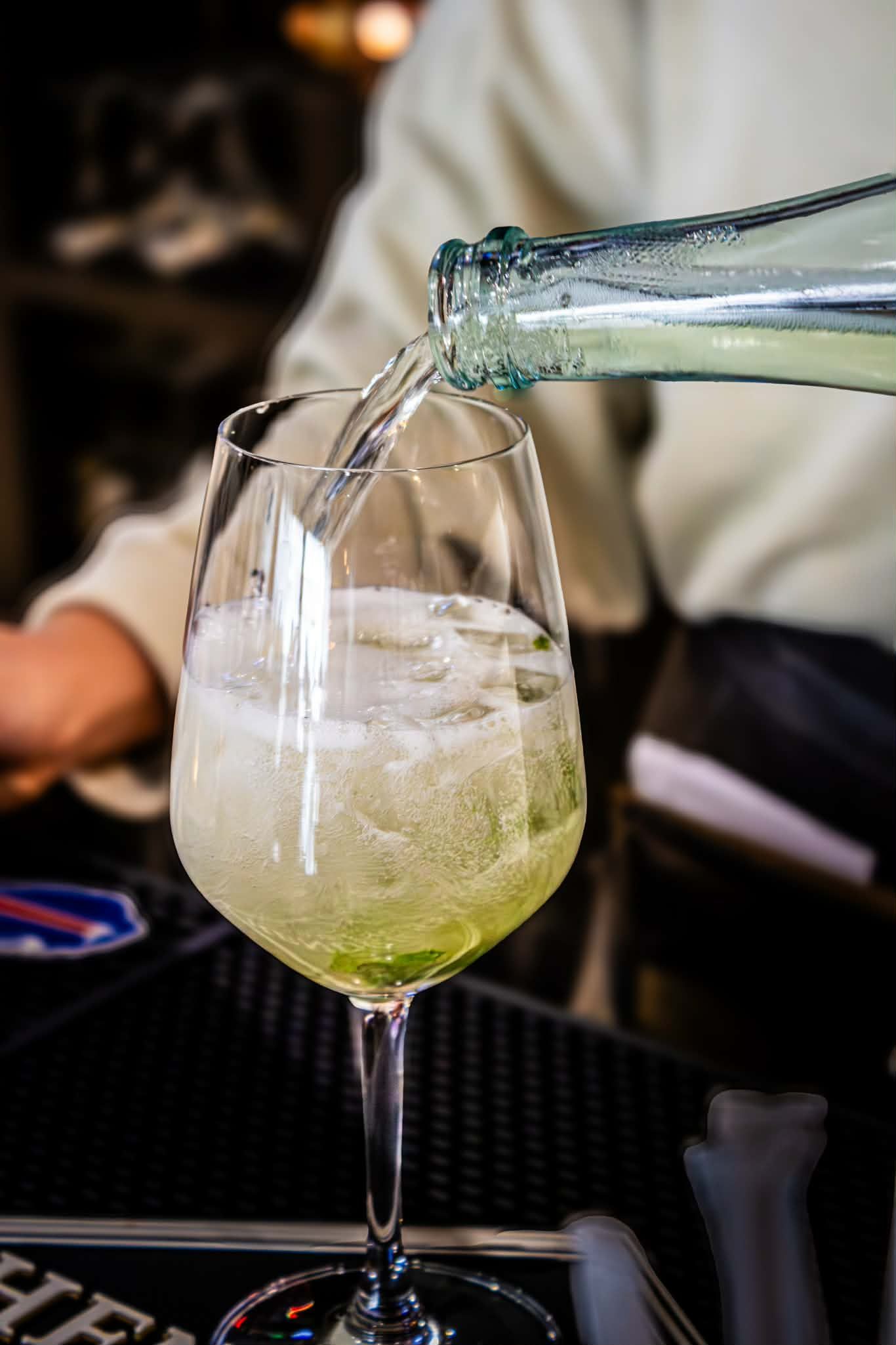 Bartender pouring sparkling white wine into a large wine glass filled with ice cubes.