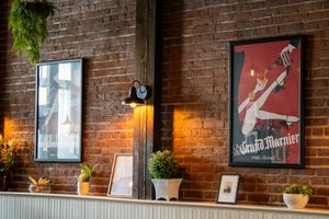 Rustic brick wall bar decor featuring Grand Marnier vintage poster, wall sconce lighting, and potted plants on a white shelf.