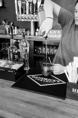 Bartender mixing a cocktail on a Hendrick's Gin bar mat, with beer taps and bottles visible in background.