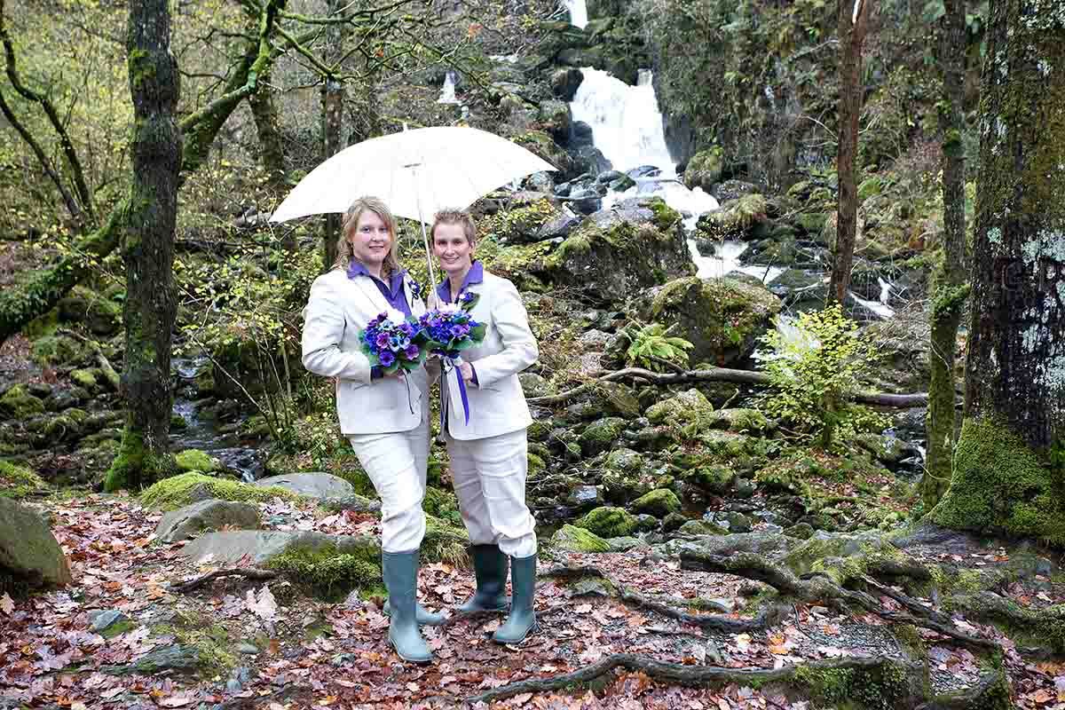 Two women in white outfits holding umbrella and purple bouquet pose by mossy waterfall in forest on their wedding day.