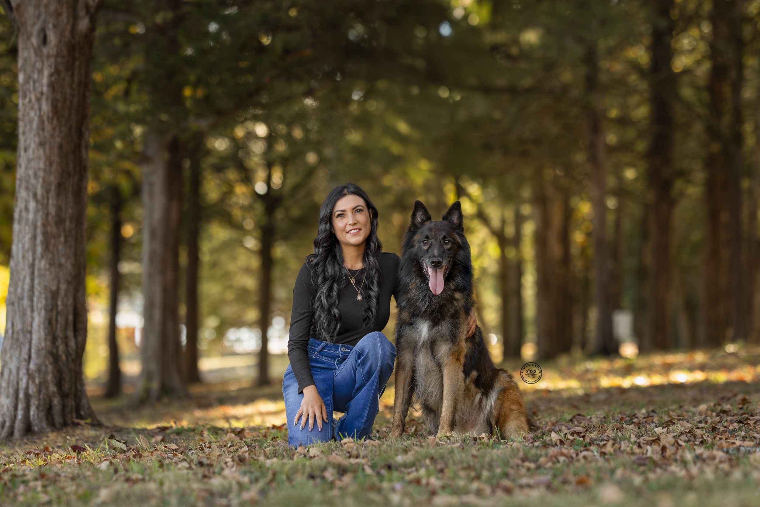 Woman kneeling beside a German Shepherd dog in a tree-lined park with autumn leaves on the ground.