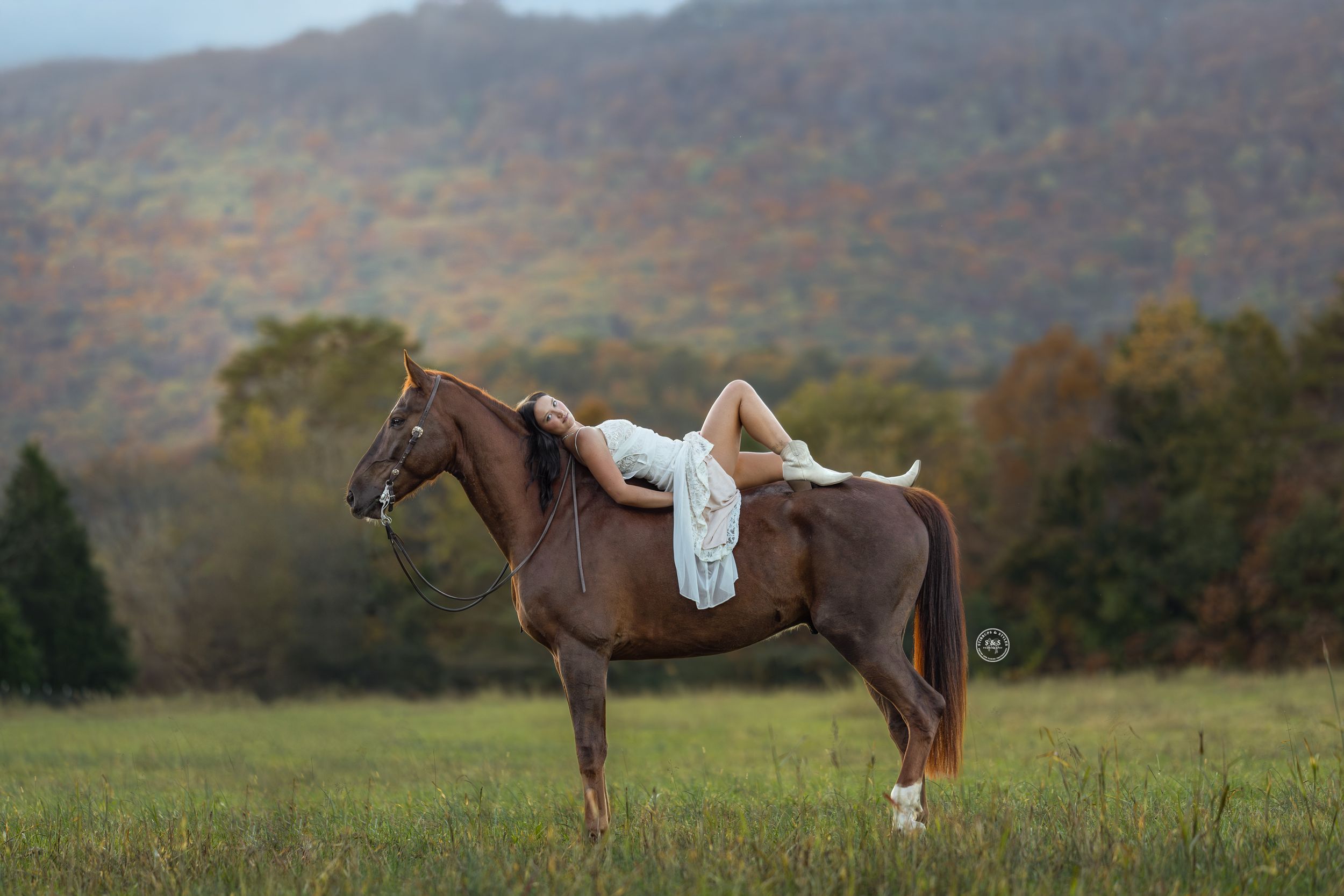 Woman in white dress lying on a chestnut horse in a green field with colorful autumn mountain backdrop.