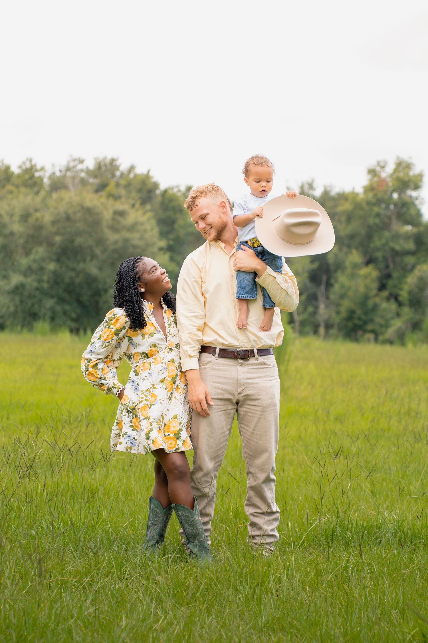 A happy interracial family poses in a green field, with a man holding a toddler wearing a cowboy hat beside a smiling woman.