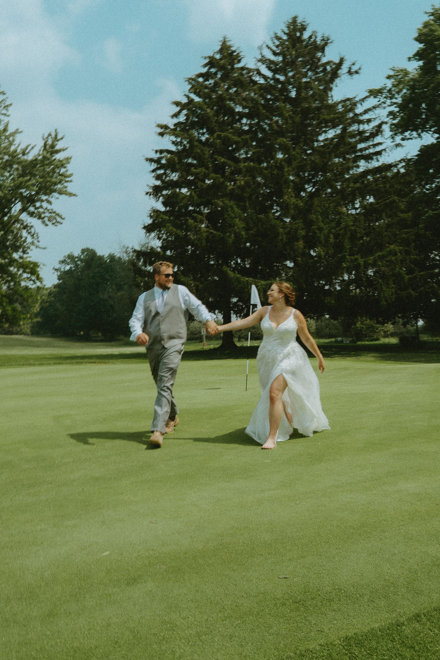 Bride and groom holding hands running on a golf course green on their wedding day with tall trees in the background.