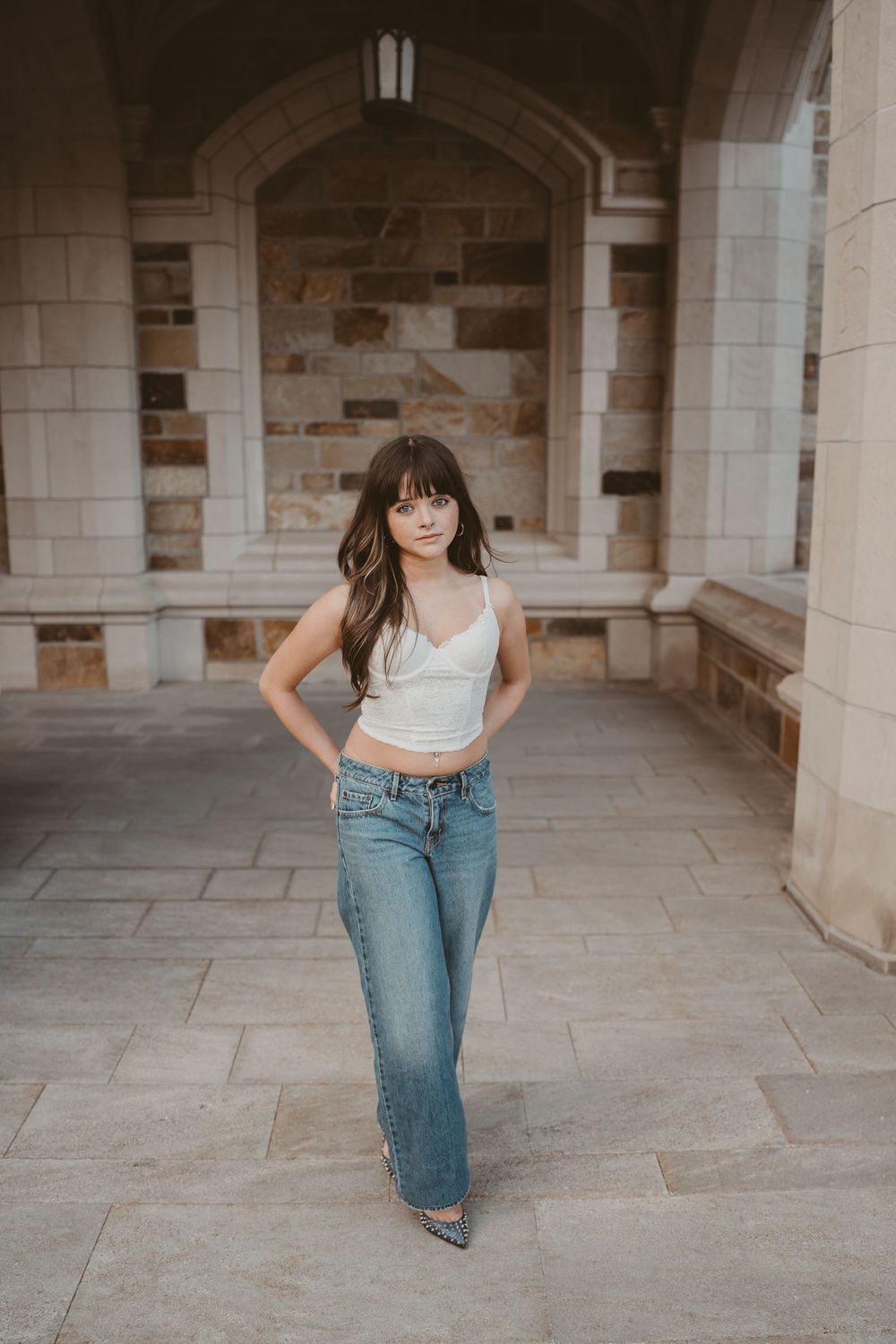 Young woman in white crop top and flared jeans poses confidently in stone archway corridor.