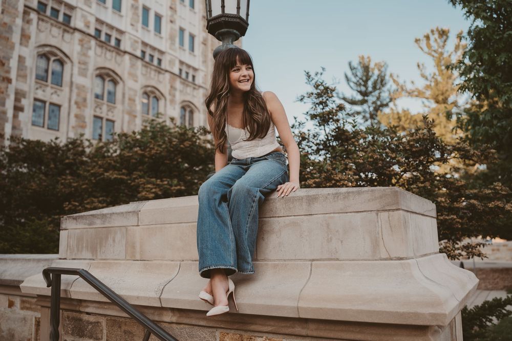 A young woman sits smiling on a stone ledge outdoors, wearing jeans and a white top, with trees and a building behind her.