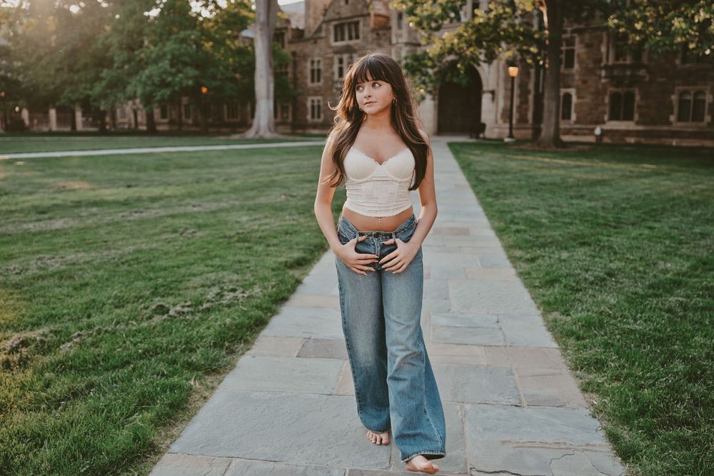 Young woman in crop top and wide-leg jeans poses on stone path in front of historic brick building.