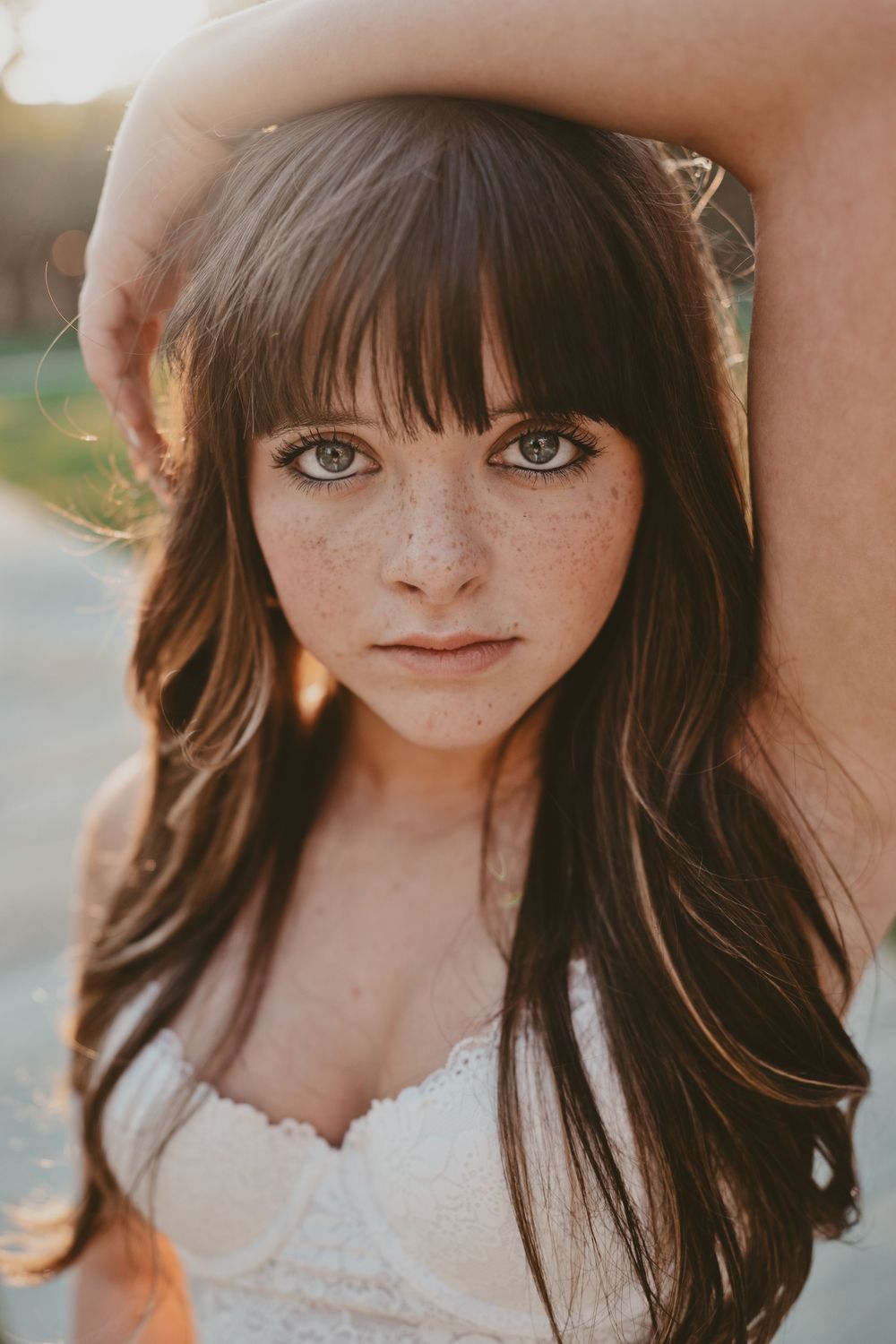 Young woman with freckles, bangs, and long brown hair poses outdoors in golden hour light wearing a white lace top.