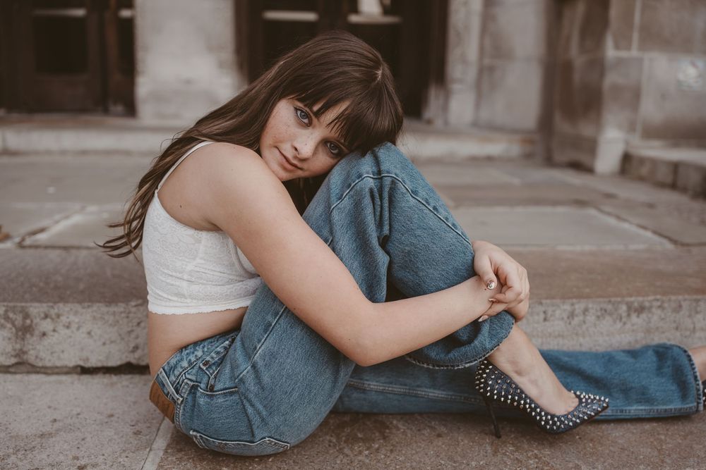 Young woman in white crop top and blue jeans sitting on steps, smiling over shoulder, wearing studded heels.