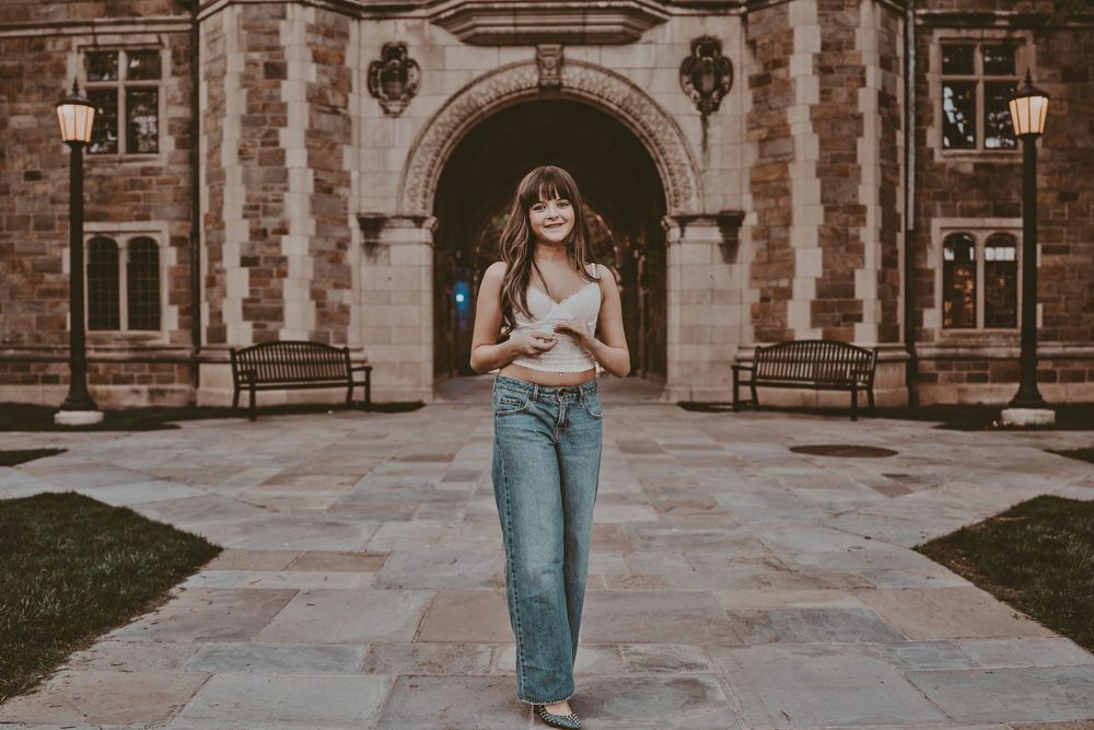 Young woman in jeans and white crop top walking in front of a historic stone university building archway.