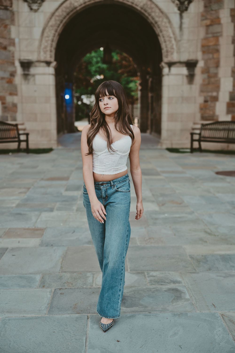 Young woman in white crop top and wide-leg jeans poses confidently on stone courtyard near historic arched building.