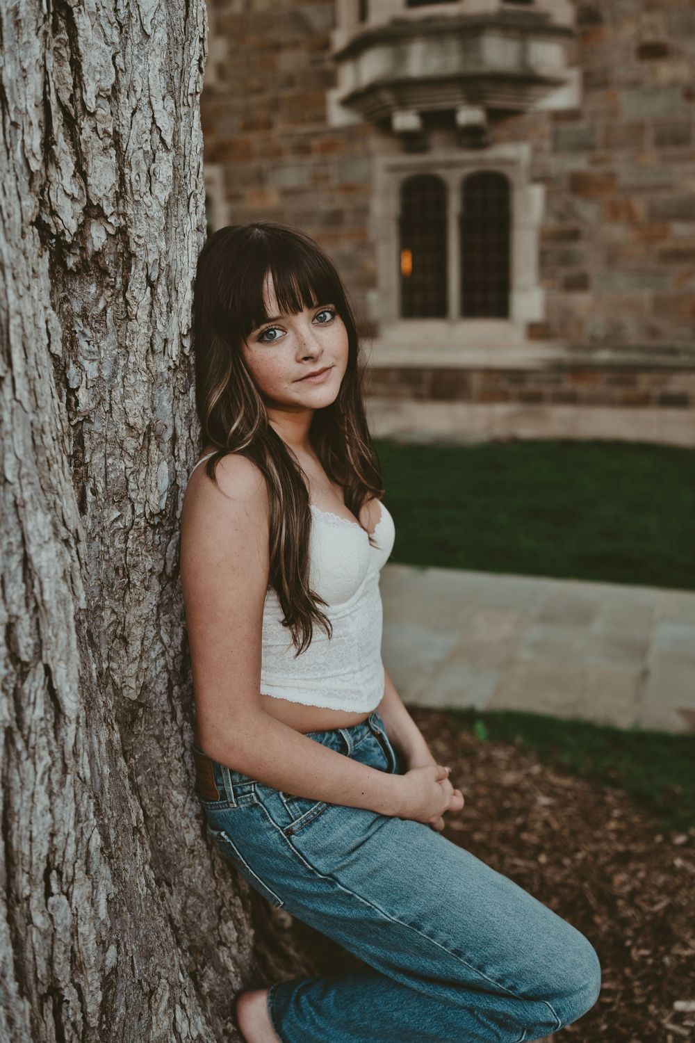 Young woman with dark hair in white tank top and jeans leaning against a tree near a stone building.