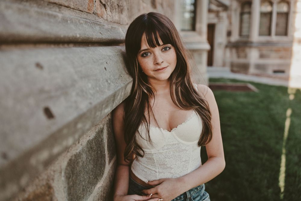 Young woman with long brown hair in white corset top leans against stone wall outdoors.