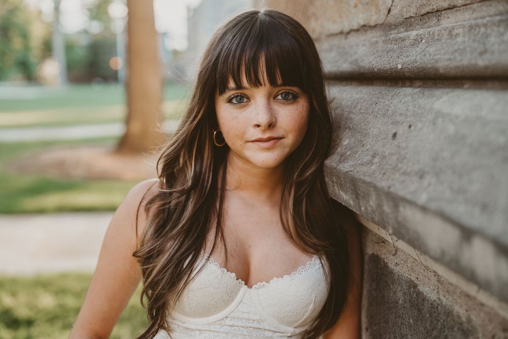 Young woman with long brunette hair and bangs leaning against a rustic stone wall outdoors.