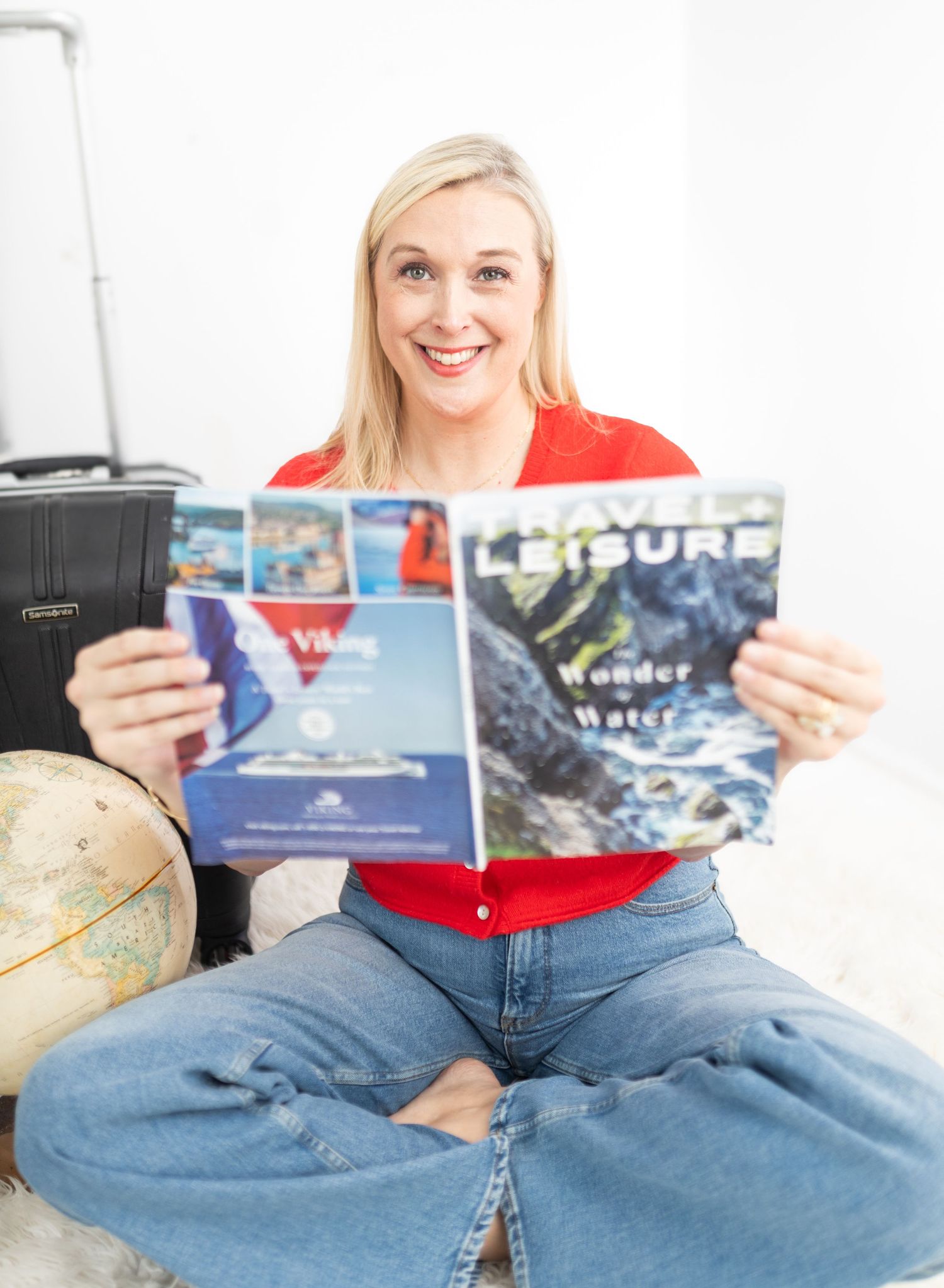 Smiling blonde woman sitting cross-legged holding a Travel Leisure magazine, with a globe and suitcase behind her.
