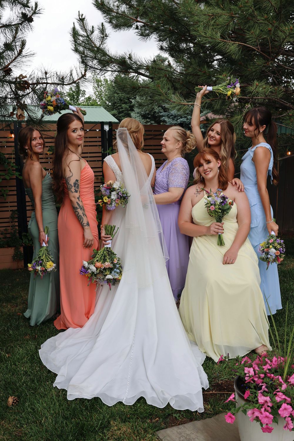 Bride in white gown with veil poses with bridesmaids in colorful pastel dresses holding floral bouquets outdoors.